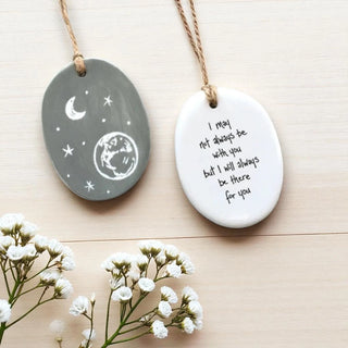 Two ceramic pendants with star and moon design and motivational quote on a wooden surface with flowers.