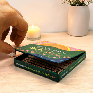 Hand opening a box of Louise Hay's Affirmations for Self-Esteem cards on a wooden surface with a candle and vase in the background.