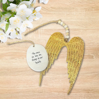 Gold angel wings with a white porcelain oval with the words the ones we love are always in our hearts, displayed on a wooden surface with flowers.