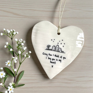Heart-shaped ceramic ornament with a house illustration and text on a wooden surface with flowers.