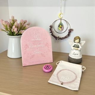 Decorative items including a pink plaque, angel figurine, suncatcher, worry stone and rose quartz beaded bracelet on a wooden surface.
