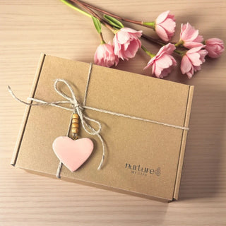 Brown gift box with pink heart charm and 'nurture' branding on a wooden surface with pink flowers.