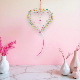 Heart-shaped decorative item with colorful beads hanging above white vases with pink flowers on a pink background.