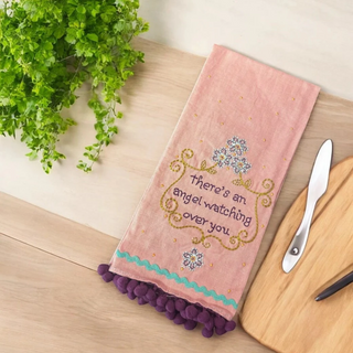 Decorative pink kitchen towel with embroidery and pom-poms on a wooden surface with a plant and knife.