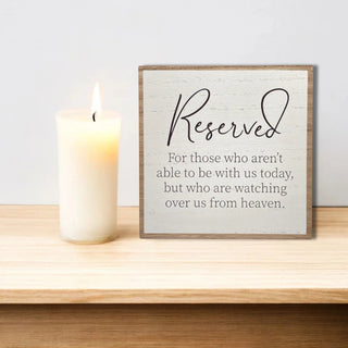 Framed sign with 'Reserved' message that says For those who aren't able to be with us today, but who are watching over us from heaven next to a lit candle on a wooden surface.