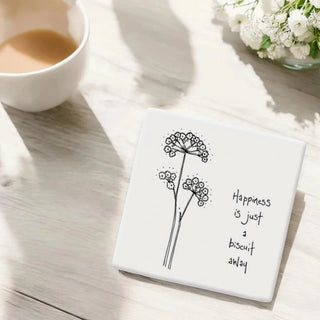 Square coaster with floral illustration and 'Happiness is just a biscuit away' text on a wooden surface with a cup of coffee and flowers.
