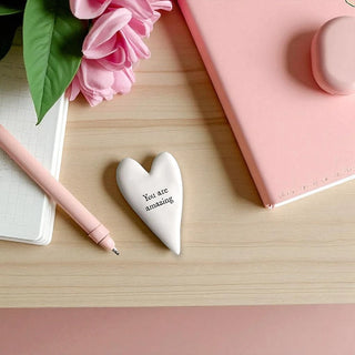 Heart-shaped white object with 'You are amazing' text on a wooden surface with pink flowers and a pen.