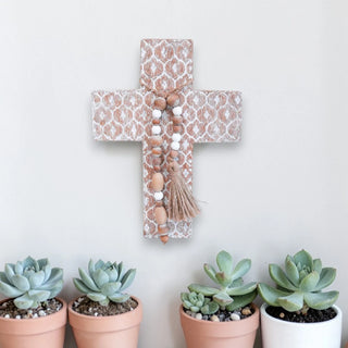 Decorative cross with beaded tassels against a white wall, with potted succulents on the floor.